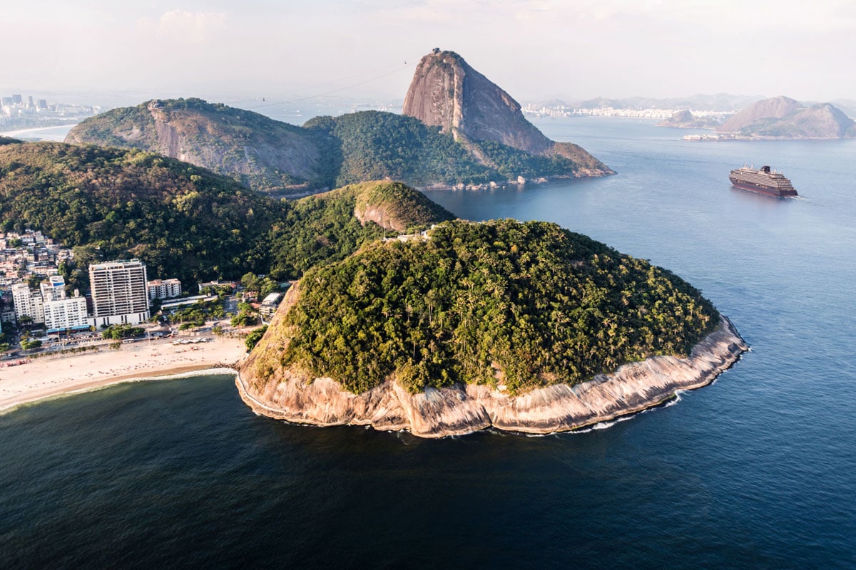 Luftaufnahme der Küste von Rio de Janeiro mit dem Zuckerhut, bewaldeten Hügeln, einem Sandstrand, städtischer Bebauung und einem Kreuzfahrtschiff im Meer.