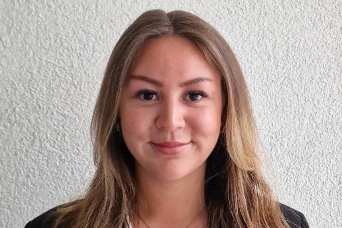 Eine junge Frau mit mittellangem, braunem Haar und freundlichem Gesichtsausdruck blickt direkt in die Kamera vor einer hellen, strukturierten Wandfläche.