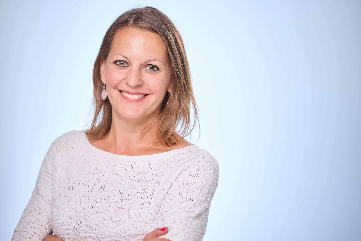 Eine lächelnde Frau mit schulterlangem, braunem Haar trägt ein weißes Oberteil mit Strukturmuster vor einem hellblauen Hintergrund im Studio-Porträt.