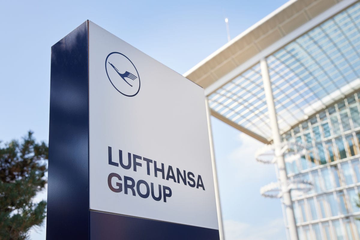 Ein vertikales Firmenschild der Lufthansa Group mit dem Kranich-Logo vor einem modernen Gebäude mit Glasfassade und Stahlkonstruktion bei blauem Himmel.