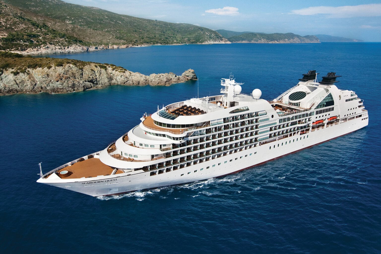 Die Seabourn Quest fährt auf dem tiefblauen Meer vor einer felsigen Küstenlandschaft mit grünen Hügeln unter einem hellen Himmel bei sonnigem Tageslicht.