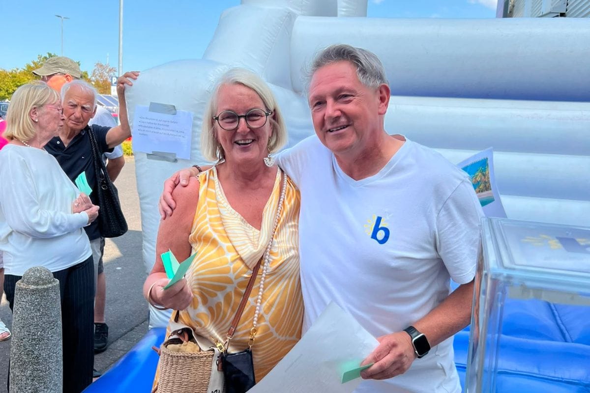 Eine Frau in gelbem Oberteil und ein Mann in weißem T-Shirt stehen lächelnd vor einer weißen Hüpfburg; im Hintergrund sind weitere Personen zu sehen.