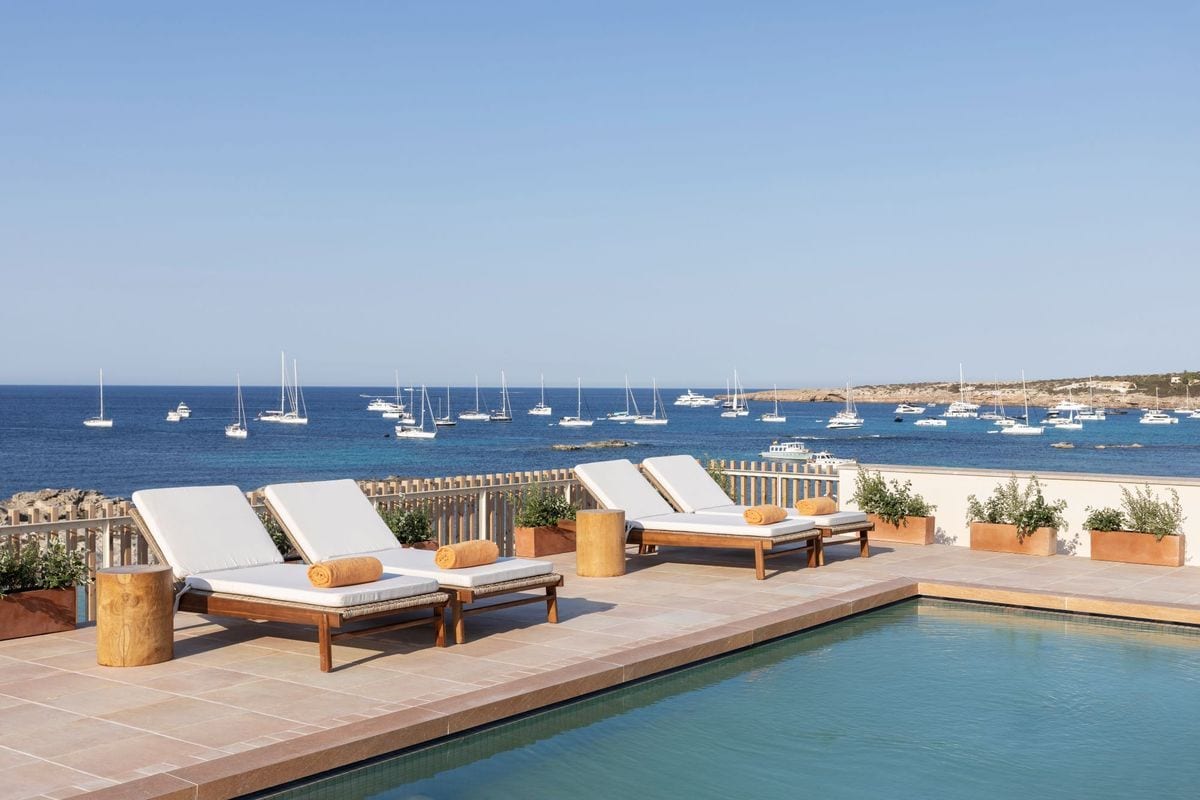 Vier weiße Sonnenliegen mit orangefarbenen Handtüchern stehen auf einer Steinterrasse an einem Pool mit Blick auf das blaue Meer mit zahlreichen Booten.