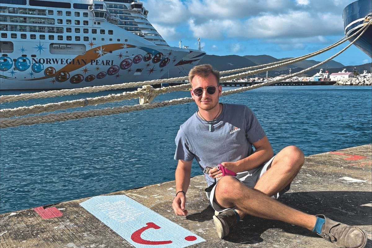 Ein junger Mann mit Sonnenbrille sitzt auf einem Kai vor der Norwegian Pearl, einem großen Kreuzfahrtschiff, das im Hafen vor einer Bergkulisse liegt.