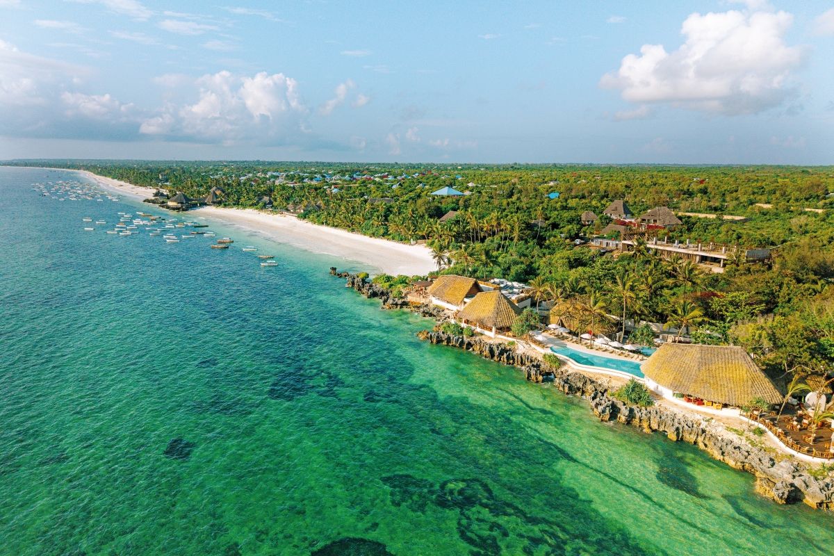 Luftaufnahme einer tropischen Küste mit türkisfarbenem Wasser, Sandstrand, kleinen Fischerbooten und einer Hotelanlage mit strohgedeckten Dächern und Pool.