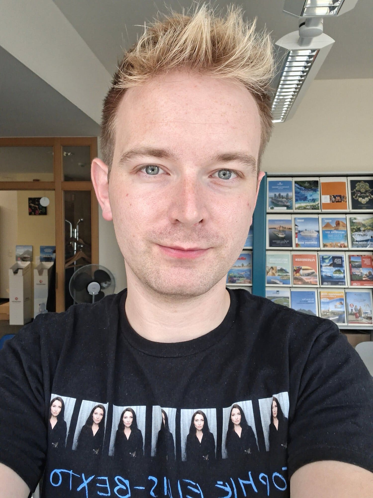 Ein junger Mann mit blondem Haar und blau-grauen Augen trägt ein schwarzes T-Shirt mit Fotodrucken vor einem Regal mit verschiedenen Reisebroschüren.