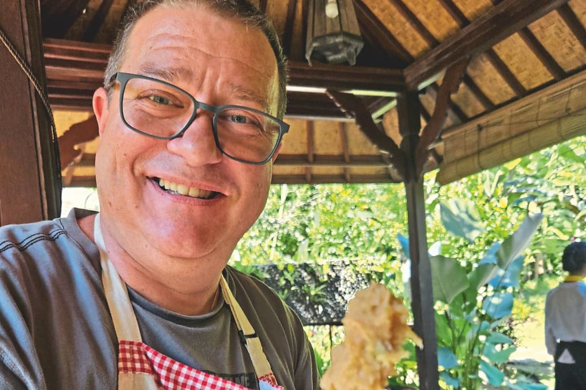 Ein lächelnder Mann mit Brille und einer rot-weiß karierten Kochschürze vor einem hölzernen Pavillon mit Blick in einen sonnigen, grünen Garten im Hintergrund.
