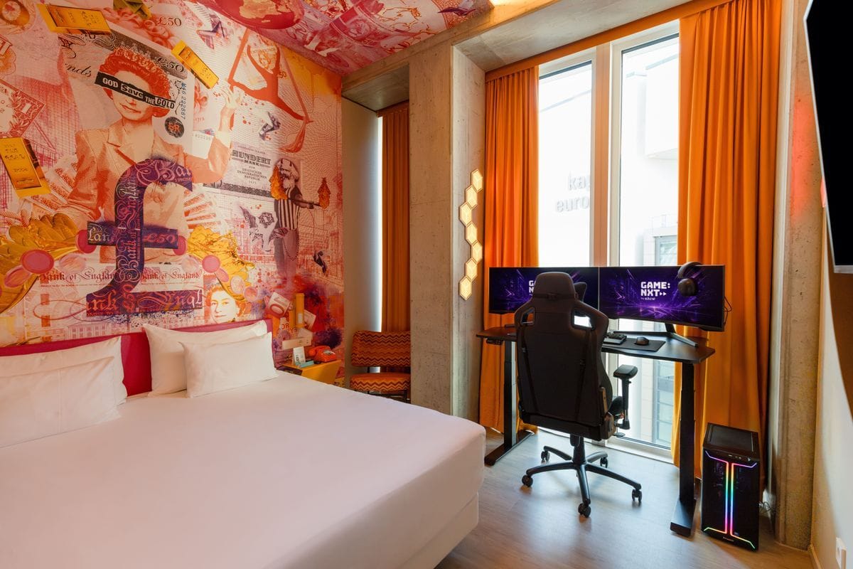 Ein Hotelzimmer mit einem Doppelbett, einer auffälligen Wandtapete mit Geldscheinmotiven und einem Gaming-Arbeitsplatz mit zwei Monitoren vor dem Fenster.