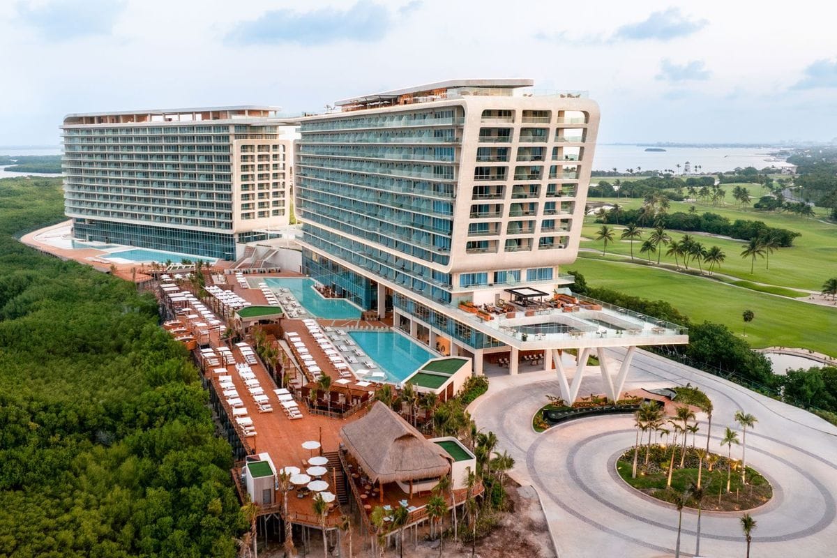 Luftaufnahme eines modernen Hotelkomplexes mit zwei großen Gebäuden, Außenpools, einer Holzterrasse mit Liegen und einer angrenzenden Golfplatzanlage.