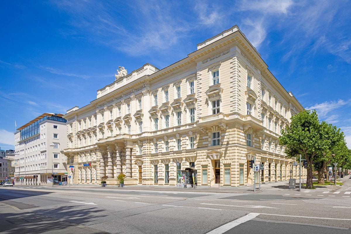 Historisches Gebäude mit eleganter Fassade und großen Fenstern, umgeben von Bäumen und modernen Gebäuden unter blauem Himmel.