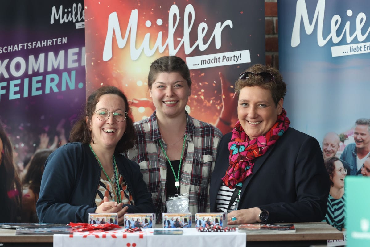 Drei lächelnde Frauen stehen hinter einem Messetisch vor Werbebannern mit der Aufschrift Müller macht Party und Meie, auf dem Infomaterialien liegen.