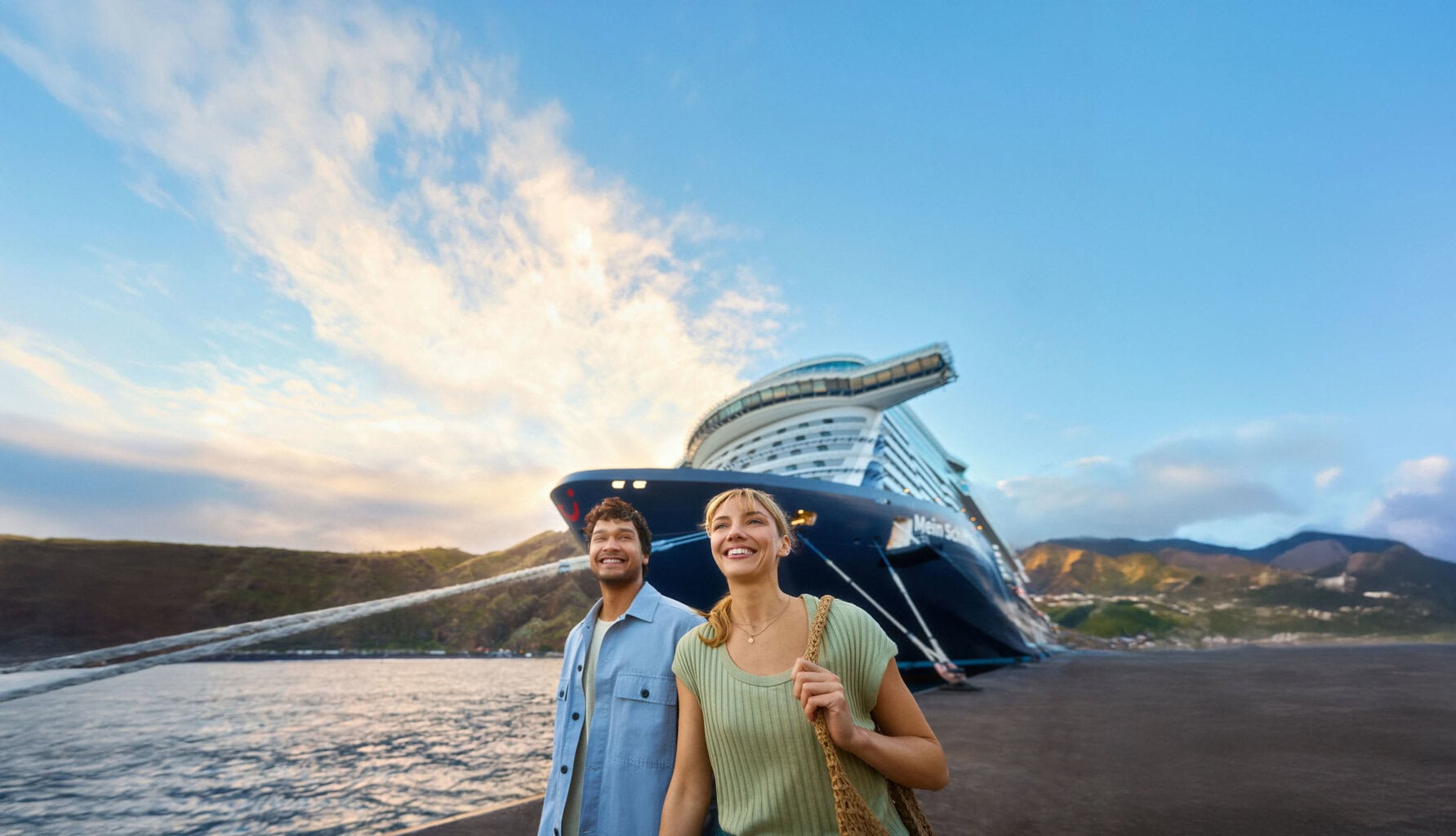 Ein junges Paar steht lächelnd auf einem Kai vor einem großen Kreuzfahrtschiff der Marke Mein Schiff, im Hintergrund sind Berge und ein blauer Himmel zu sehen.