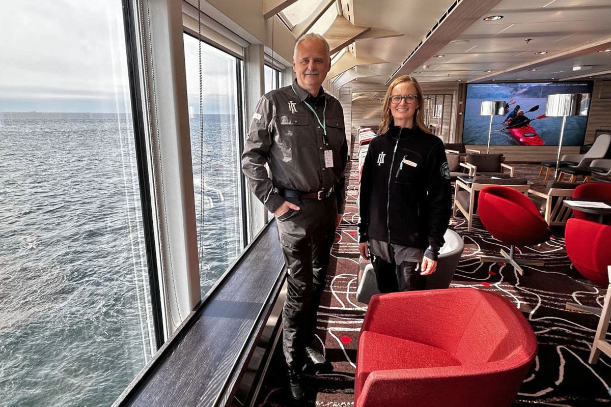 Ein Mann und eine Frau stehen in einer modernen Lounge mit großen Panoramafenstern und Blick auf das Meer, im Hintergrund sind rote Sessel zu sehen.