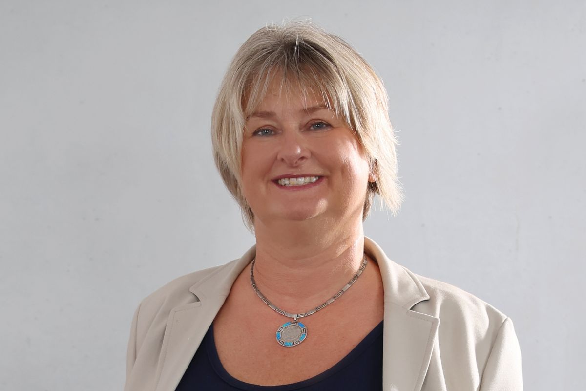 Eine Frau mit blondem, schulterlangem Haar lächelt in die Kamera. Sie trägt ein beiges Sakko über einem dunklen Oberteil und eine auffällige Halskette.