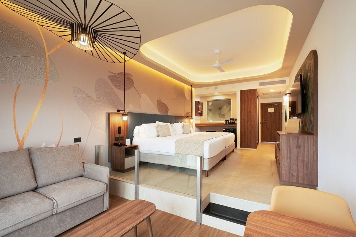 Ein modernes Hotelzimmer mit einem leicht erhöhten Schlafbereich, einem Doppelbett, einem grauen Sofa im Vordergrund und einer auffälligen Deckenleuchte.