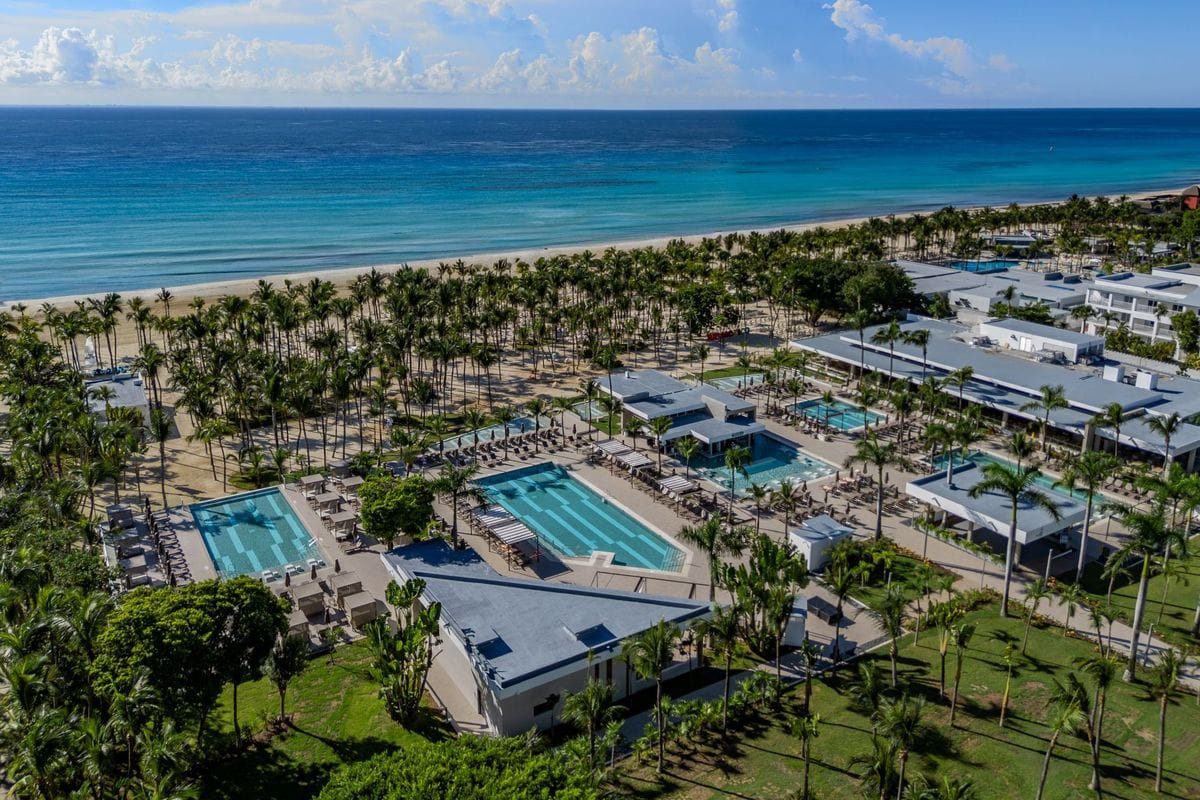 Luftaufnahme einer weitläufigen Hotelanlage mit mehreren Swimmingpools, Palmen und direktem Zugang zu einem breiten Sandstrand am türkisfarbenen Meer.