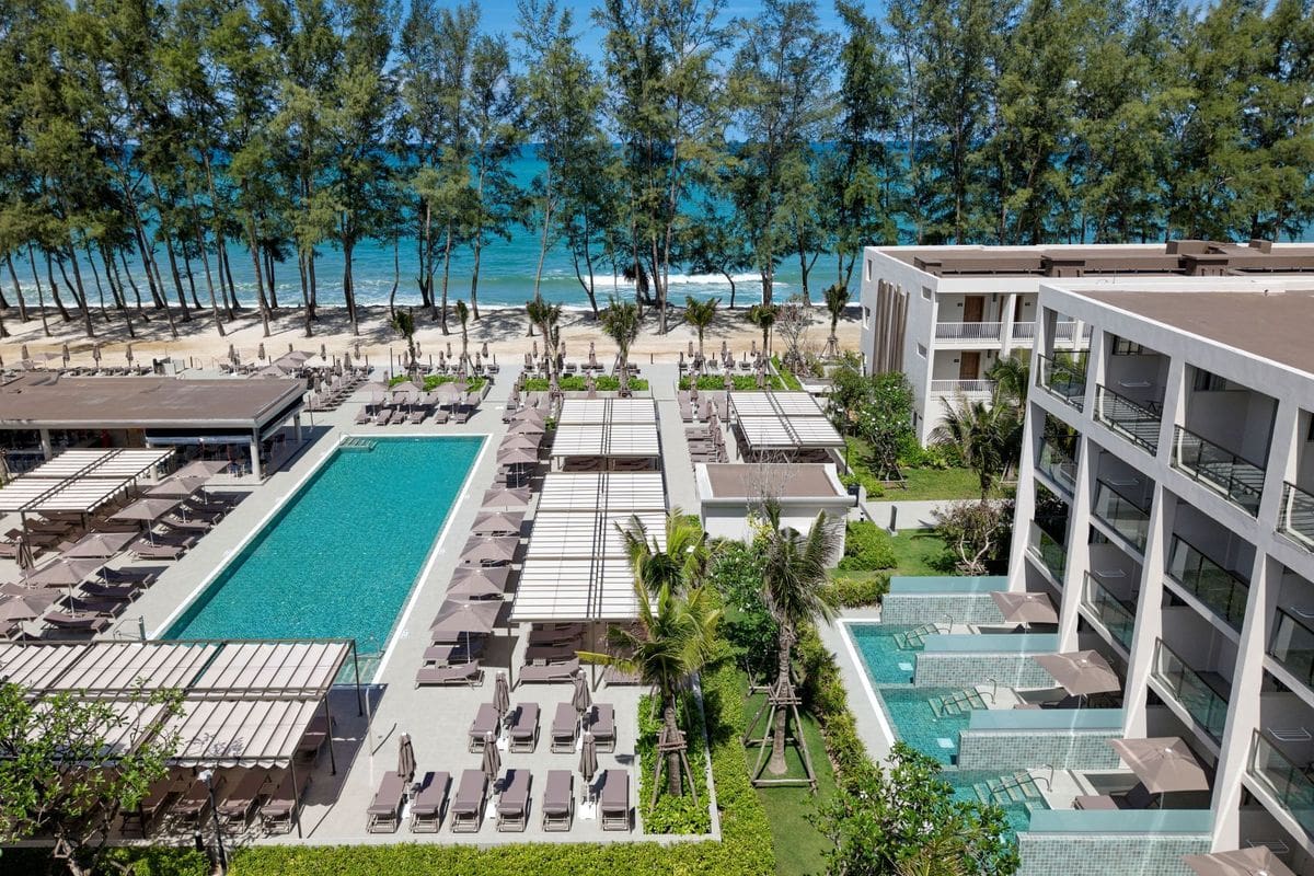 Luftaufnahme einer Hotelanlage mit einem langen rechteckigen Pool, zahlreichen Sonnenliegen, Palmen und direktem Zugang zu einem Sandstrand mit Meerblick.