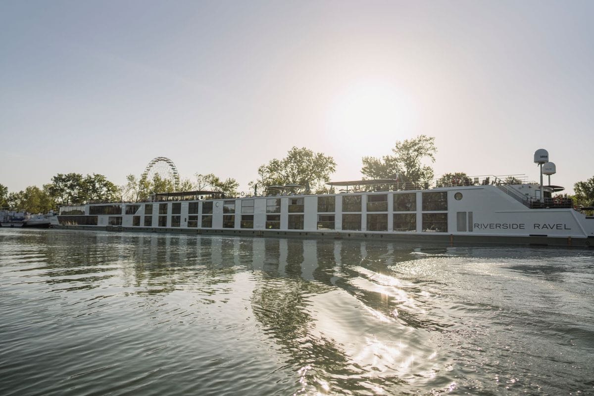 Das weiße Flusskreuzfahrtschiff Riverside Ravel fährt auf einem Fluss, im Hintergrund sind Bäume und ein Riesenrad bei hellem Sonnenlicht zu sehen.