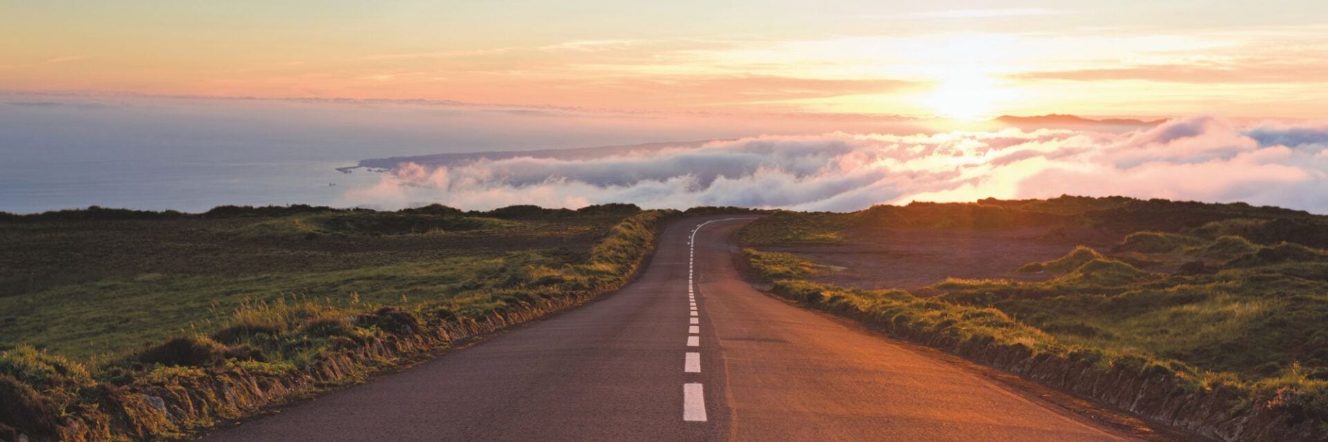 roadtrips Eine asphaltierte Straße führt durch eine hügelige, grüne Landschaft auf ein Meer aus Wolken zu, das im warmen Licht des Sonnenuntergangs erstrahlt.