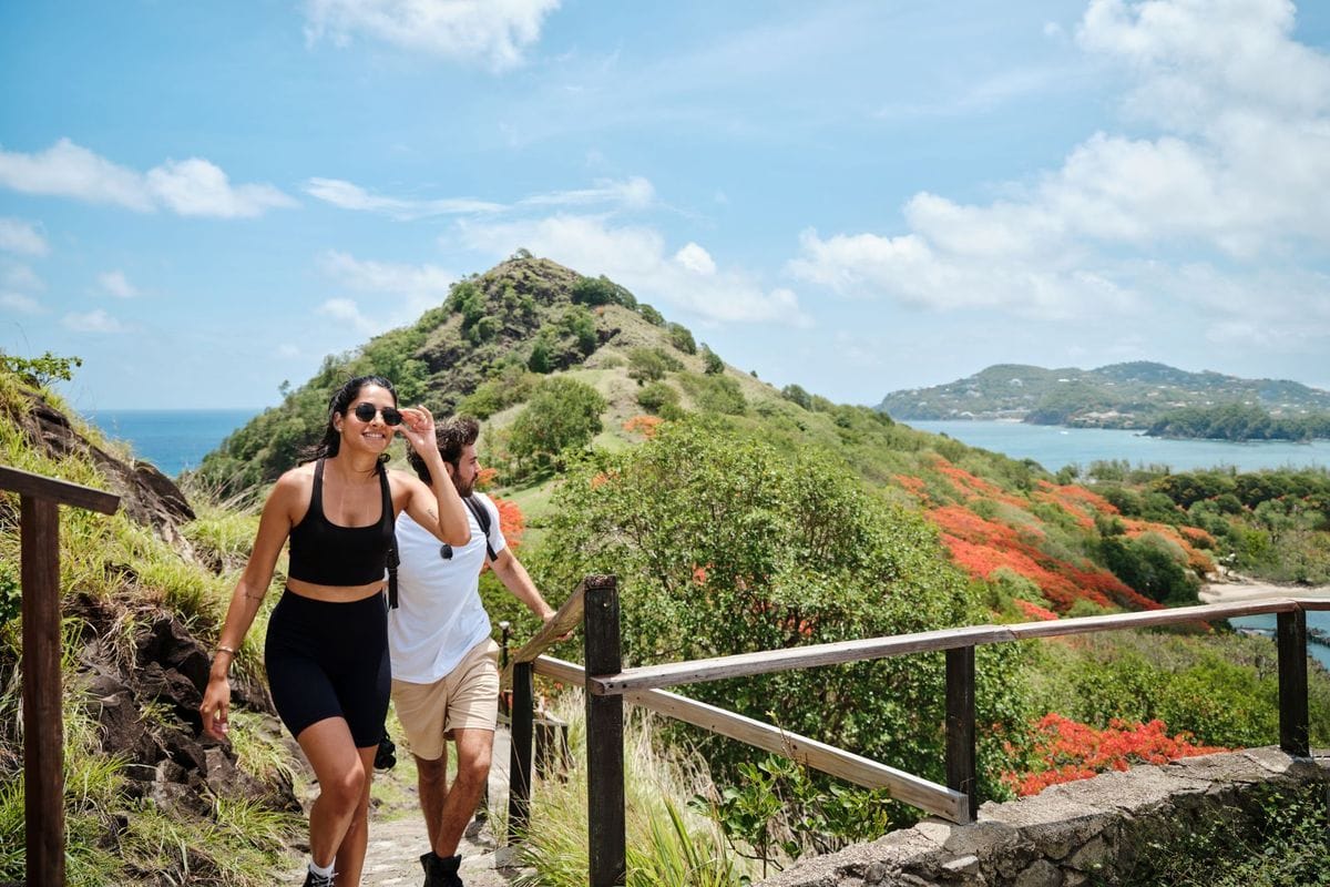 Zwei Personen wandern auf einem Pfad mit Blick auf eine grüne Landschaft und das Meer im Hintergrund, umgeben von bunten Blumen.
