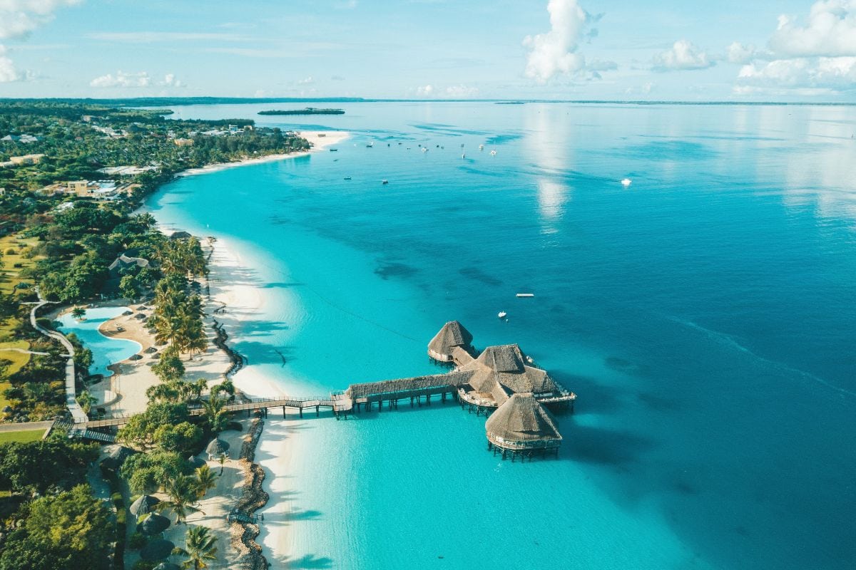 Luftaufnahme einer tropischen Küste mit weißem Sandstrand, Palmen, einem Pool und einem langen Holzsteg, der zu strohgedeckten Stelzenhäusern im Meer führt.