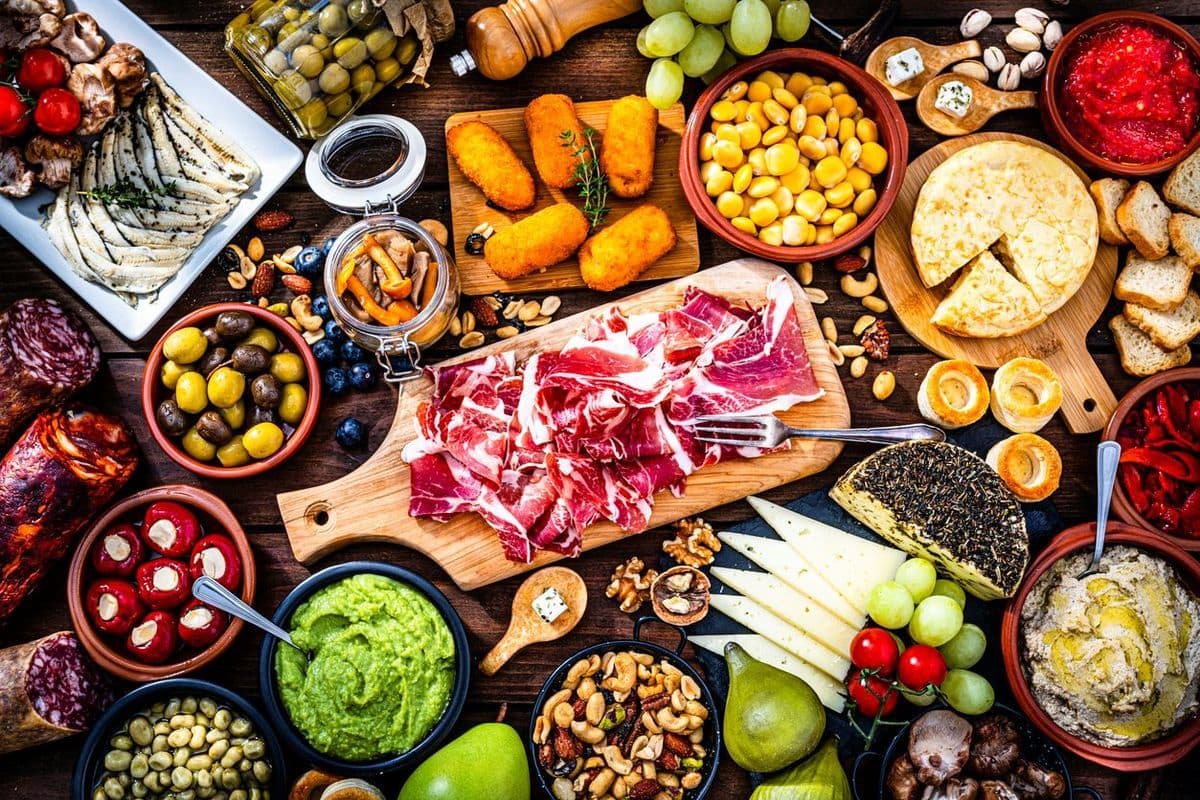 Draufsicht auf einen rustikalen Holztisch mit einer vielfältigen Auswahl an Tapas, darunter Schinken, Käse, Oliven, Kroketten, Dips und frisches Obst.