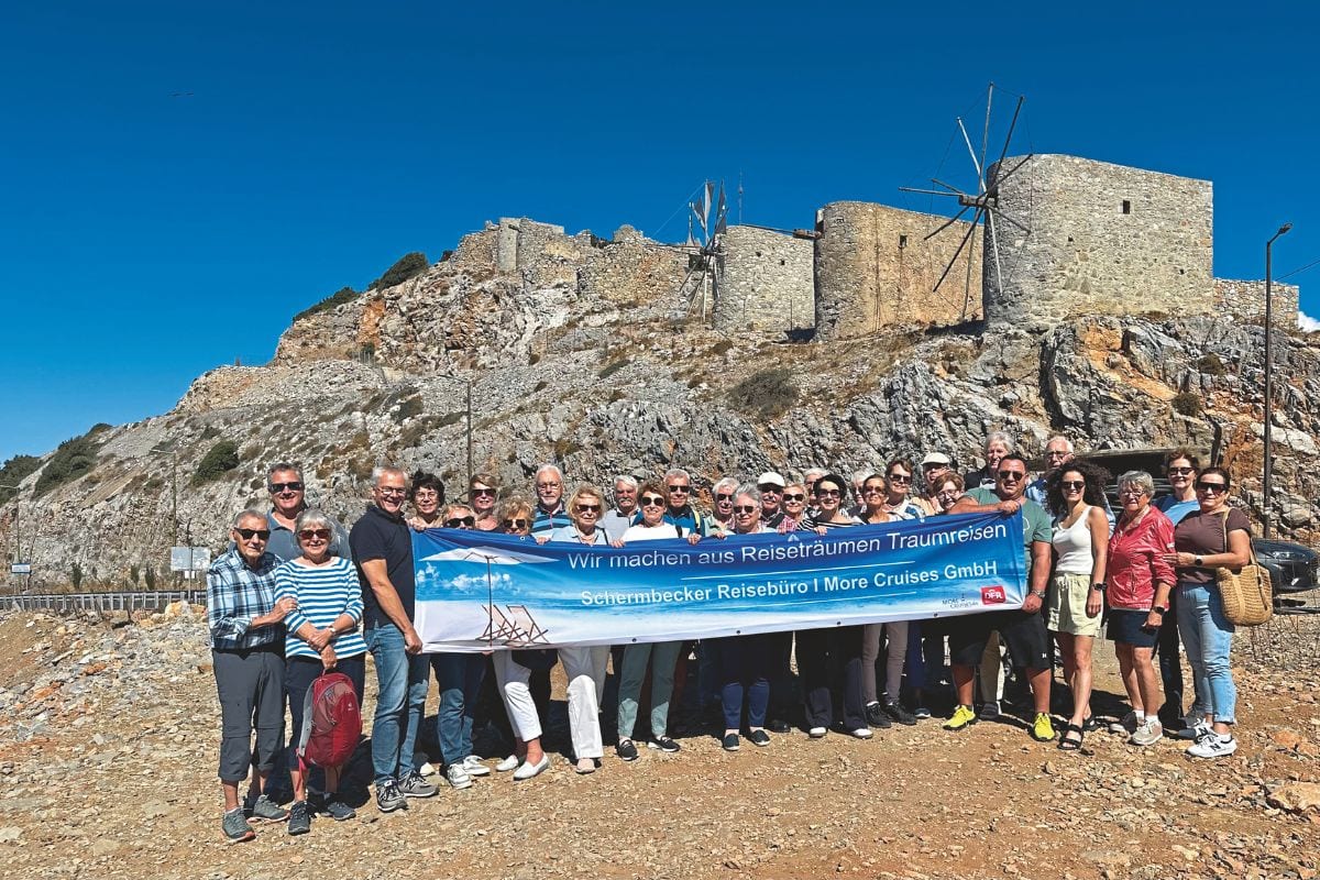 Eine große Gruppe von Menschen hält gemeinsam ein blaues Banner vor einer felsigen Hügellandschaft mit alten Windmühlen unter einem klaren blauen Himmel.