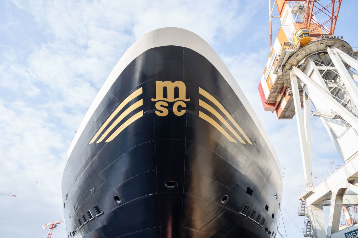 Die Frontansicht des schwarzen Bugs eines MSC-Kreuzfahrtschiffes mit goldenem Logo vor einem hellblauen Himmel und einem großen weißen Werftkran im Hintergrund.