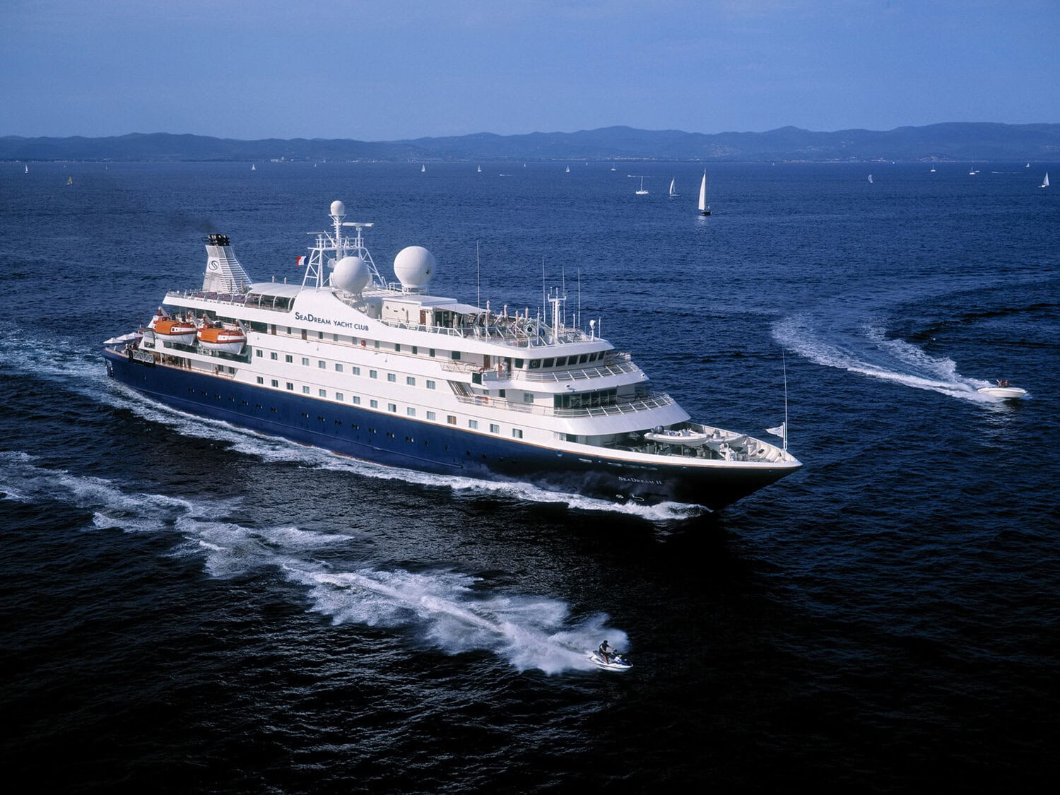 Die SeaDream II fährt auf dem offenen Meer, begleitet von einem Jetski im Vordergrund und weiteren Segelbooten in der Ferne vor einer hügeligen Küstenlinie.