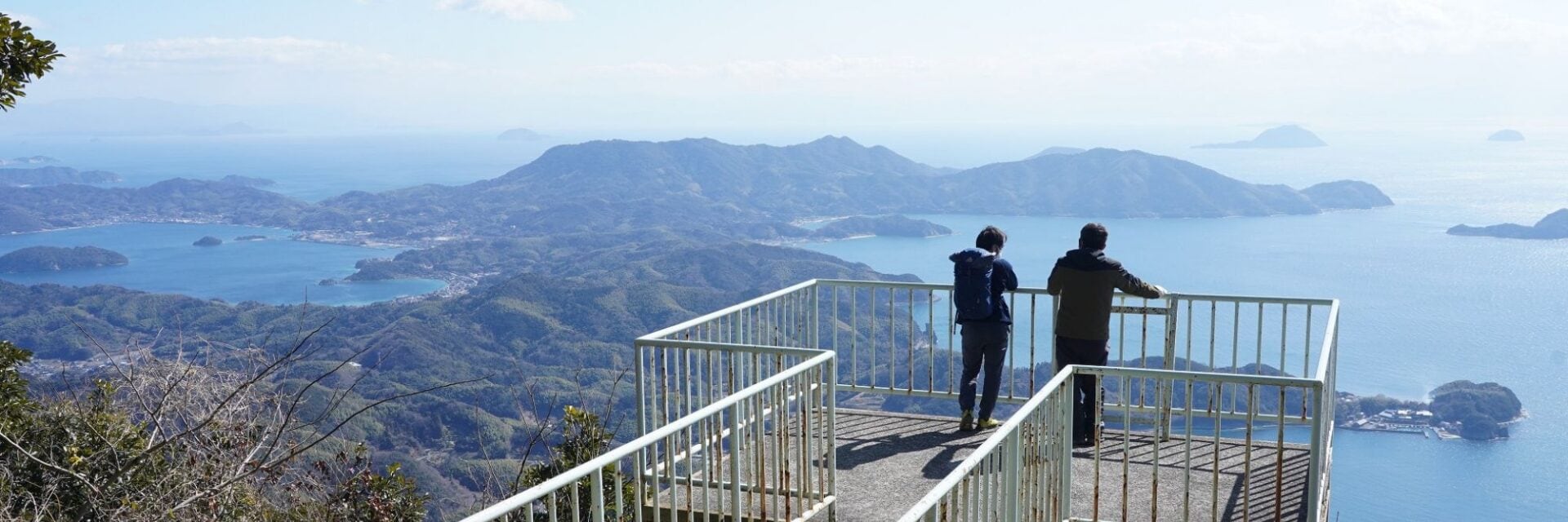 setouchi alps trekking hero Zwei Personen stehen auf einer erhöhten Aussichtsplattform mit weißem Metallgeländer und blicken auf eine weitläufige Küstenlandschaft mit Inseln und Meer.