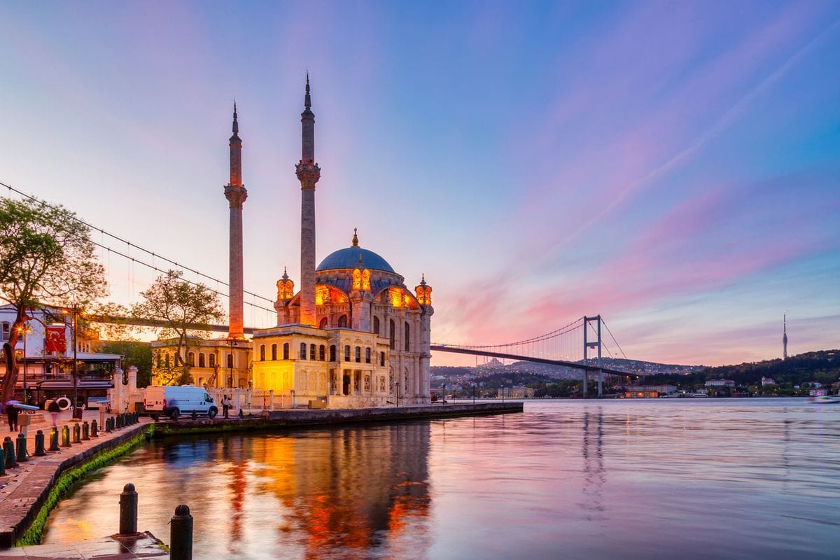 Die Ortaköy-Moschee in Istanbul bei Sonnenuntergang mit ihren zwei Minaretten am Ufer des Bosporus, im Hintergrund die beleuchtete Bosporus-Brücke am Abend.