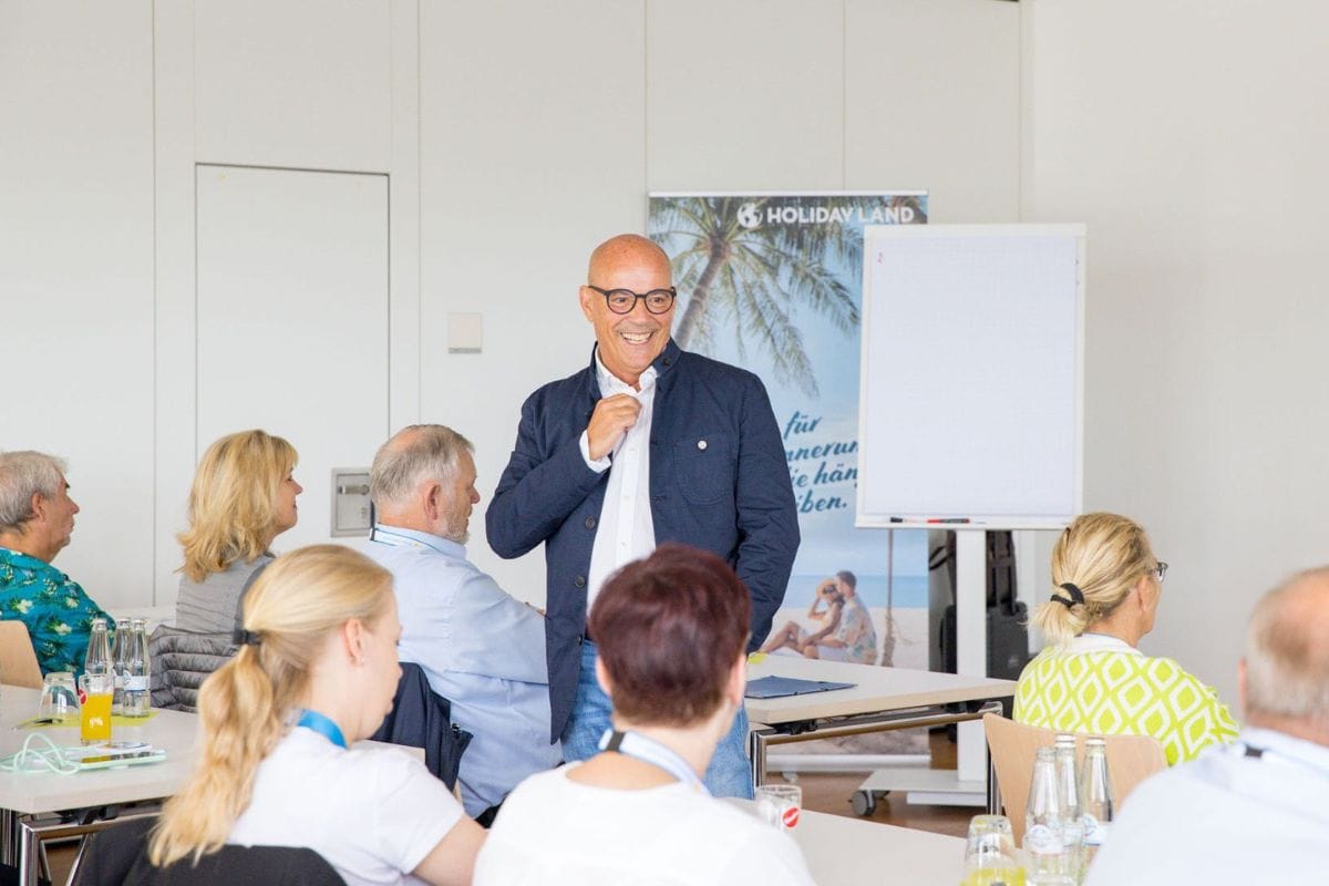 Ein lächelnder Mann mit Brille und dunkelblauem Sakko steht vor einer Gruppe von Menschen in einem hellen Konferenzraum neben einem Holiday Land Banner.