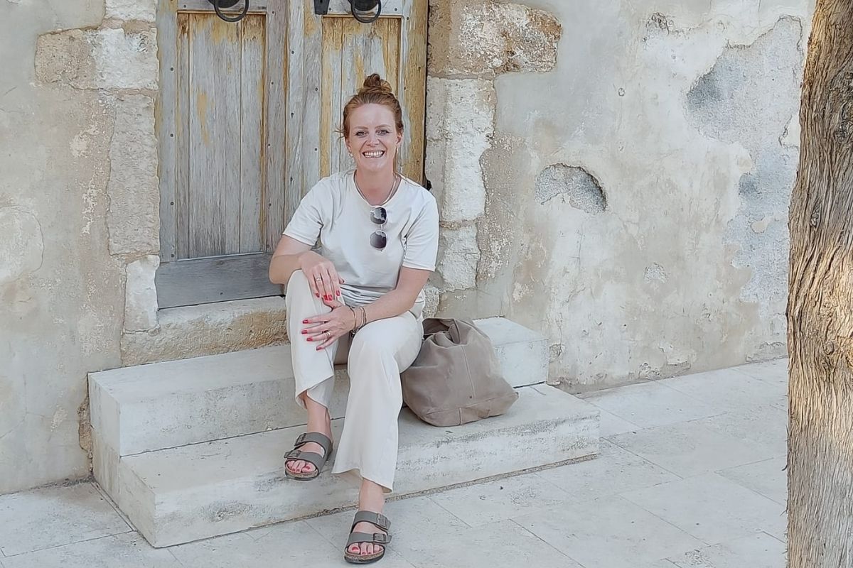 Eine lächelnde Frau mit hochgesteckten Haaren sitzt in heller Kleidung auf einer Steinstufe vor einer alten Holztür mit einer beigefarbenen Tasche daneben.
