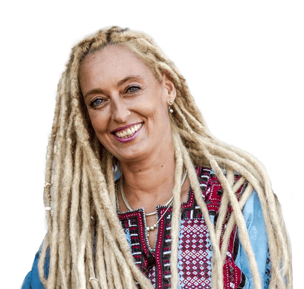 stella-deetjen- Eine lächelnde Frau mit langen, hellblonden Dreadlocks trägt eine gemusterte, farbenfrohe Bluse und eine silberne Halskette vor einem hellblauen Hintergrund.