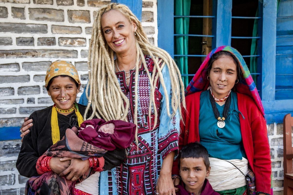 Eine Frau mit blonden Dreadlocks und gemustertem Kleid steht lächelnd neben zwei Frauen in traditioneller Kleidung, die ein Baby und einen Jungen halten.