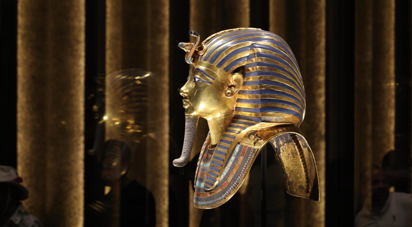 Die goldene Totenmaske des Tutanchamun in der Seitenansicht, präsentiert in einer beleuchteten Vitrine vor einem dunklen Hintergrund mit vertikalen Säulen.