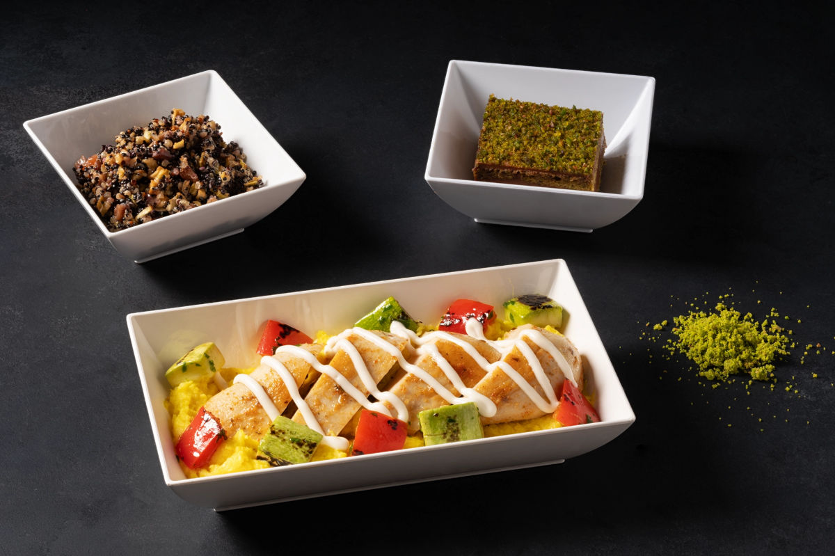Ein Teller mit gegrilltem Hähnchen, buntem Gemüse und einer weißen Sauce, begleitet von zwei Schalen mit Salat und einem Dessert.