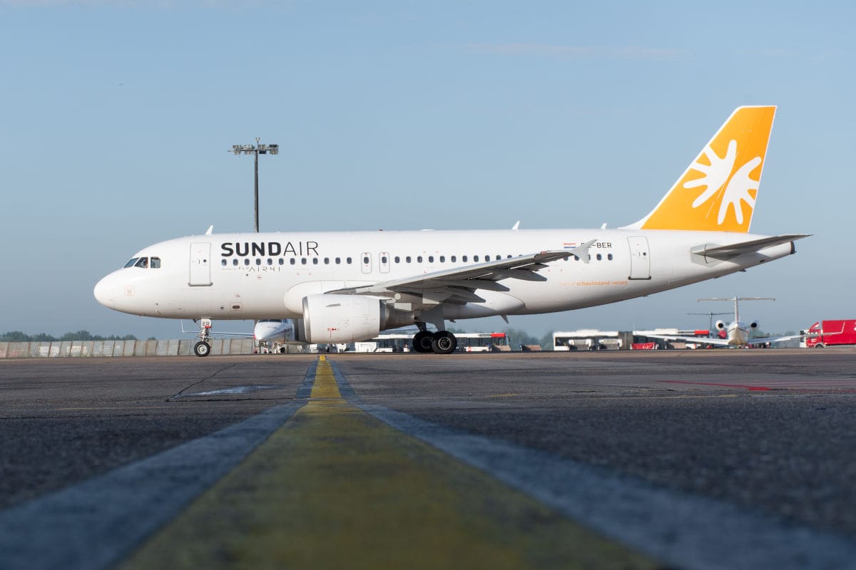 Ein weißes Passagierflugzeug der Fluggesellschaft Sundair mit gelbem Leitwerk steht auf dem Rollfeld eines Flughafens bei klarem Tageslicht zur Ansicht.