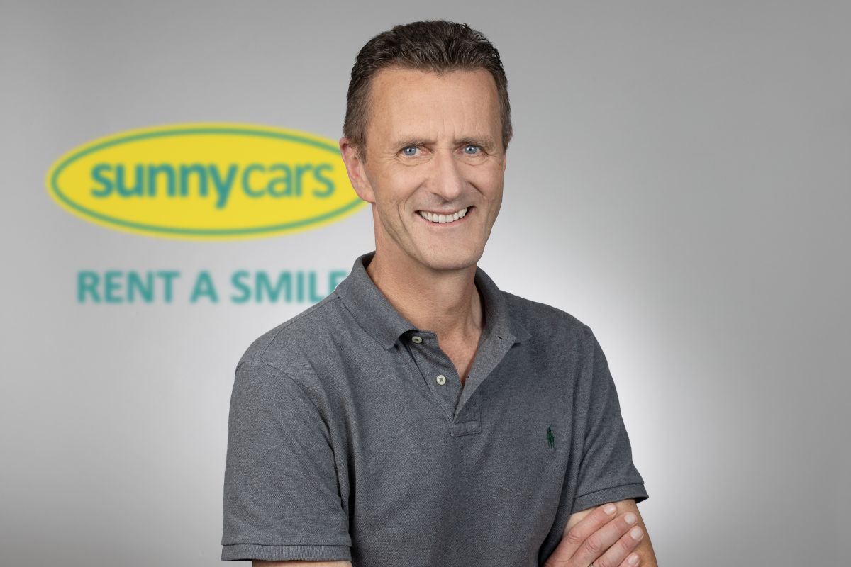 Ein lächelnder Mann mit kurzem Haar trägt ein graues Polohemd und steht vor einem unscharfen Hintergrund mit dem Logo von Sunny Cars in Gelb und Grün.