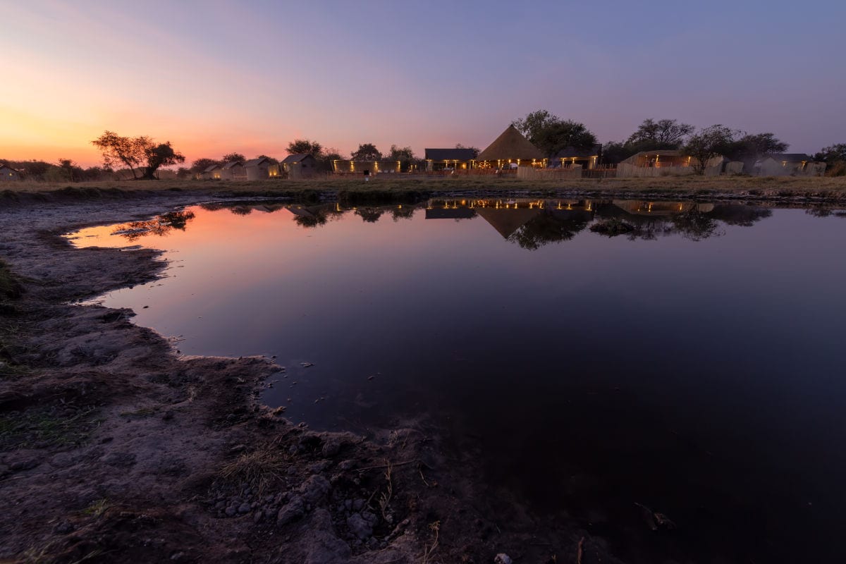 Eine Lodge mit strohgedeckten Gebäuden spiegelt sich in einem ruhigen Wasserloch bei Sonnenuntergang in einer weitläufigen, flachen Savannenlandschaft wider.