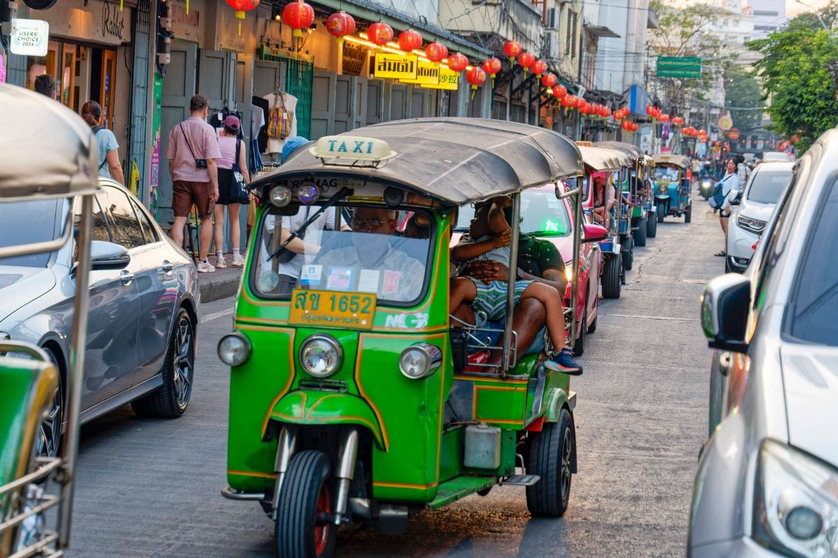 Ein grünes Tuk-Tuk mit Fahrgästen fährt auf einer belebten Straße in Bangkok, umgeben von weiteren Autos und einer Reihe roter Lampions über der Fahrbahn.