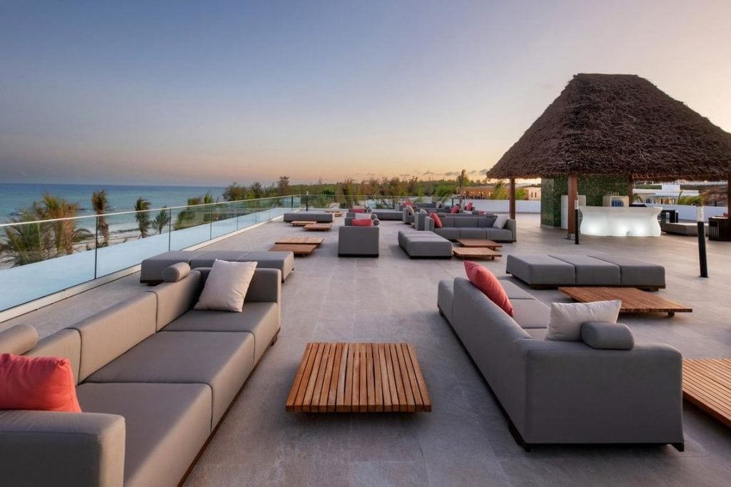 Eine weitläufige Dachterrasse mit grauen Loungemöbeln, Holztischen und einer strohgedeckten Bar bei Sonnenuntergang mit Blick auf das nahegelegene Meer.