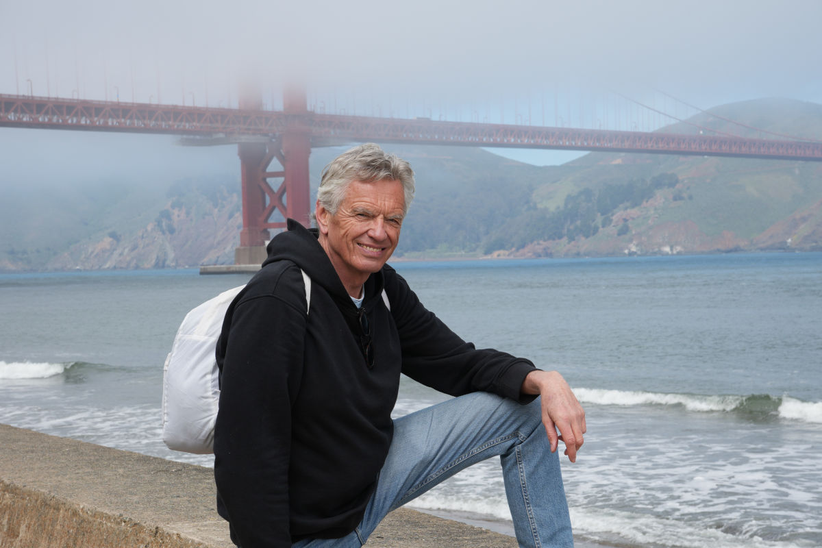 Ein älterer Mann mit grauem Haar und schwarzem Kapuzenpullover sitzt an der Küste vor der Golden Gate Bridge in San Francisco, die teilweise im Nebel liegt.
