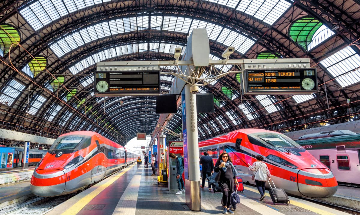 Zwei moderne Züge am Bahnhof Milano Centrale unter einem beeindruckenden Glasdach, mit Reisenden und Anzeigetafeln im Hintergrund.