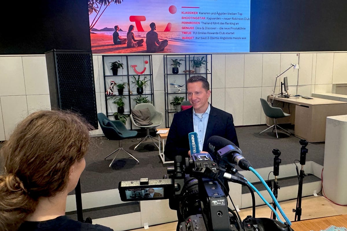 Ein Mann im dunklen Sakko wird von einer Kamerafrau interviewt. Im Hintergrund steht ein Regal mit Pflanzen vor einer Leinwand mit TUI-Logo und Text.