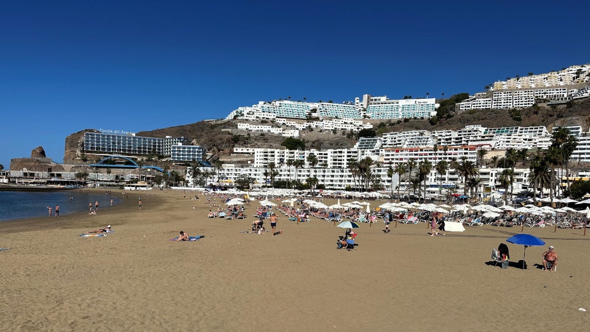 Ein breiter Sandstrand mit zahlreichen Sonnenschirmen und Liegen vor einer terrassenförmig am Hang erbauten Hotelanlage unter einem klaren, blauen Himmel.