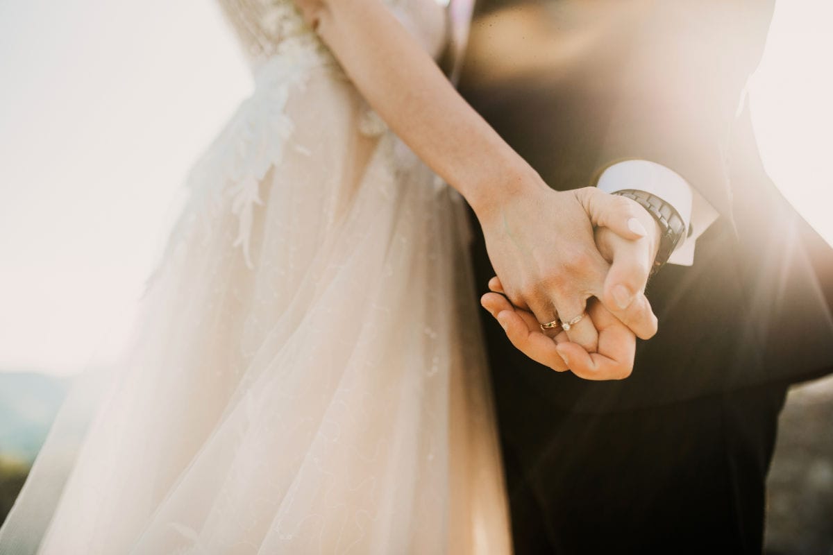Nahaufnahme eines Paares, das sich an den Händen hält. Die Person links trägt ein helles Spitzenkleid, die Person rechts einen dunklen Anzug mit Armbanduhr.