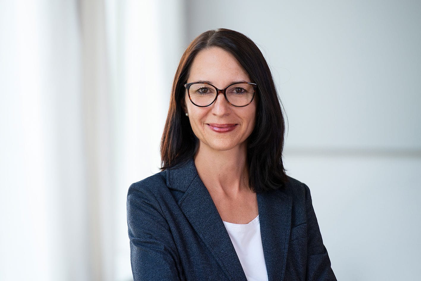 Eine Frau mit Brille und dunklem Blazer lächelt freundlich in die Kamera, vor einem hellen Hintergrund.
