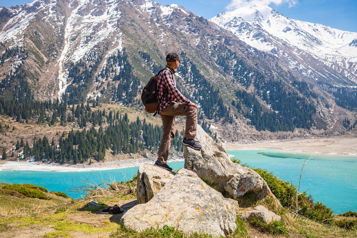 Ein Wanderer mit Rucksack steht auf einem Felsen vor einem türkisfarbenen Gebirgssee und schneebedeckten Gipfeln in einer weitläufigen Hochgebirgslandschaft.