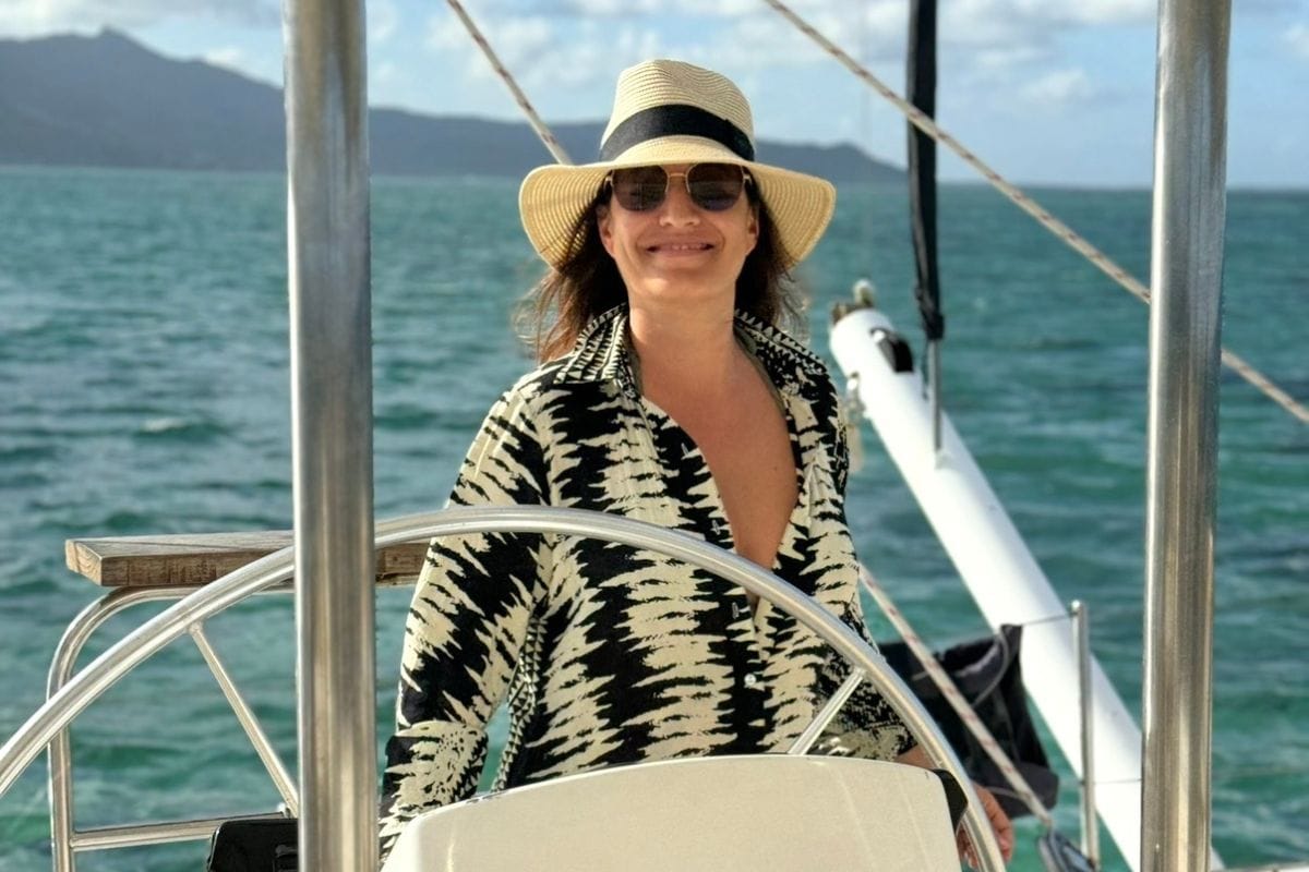 Eine Frau mit Sonnenhut und Sonnenbrille steht am Steuer eines Segelboots auf dem offenen Meer, im Hintergrund sind Berge unter einem blauen Himmel zu sehen.